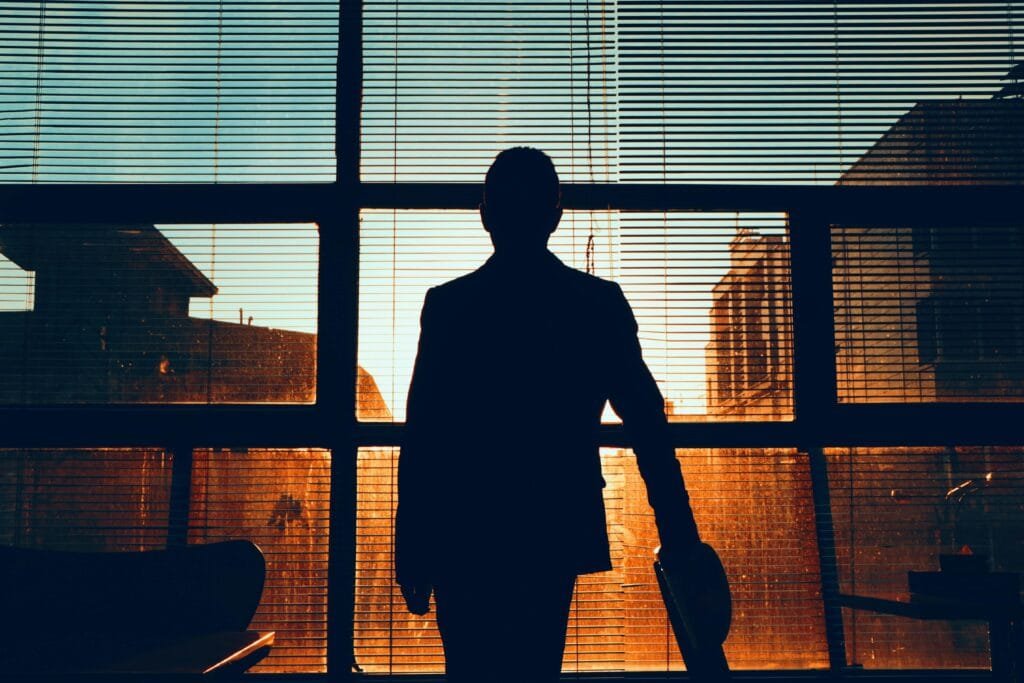 pexels-photo-7540928-7540928 Silhouette of a man standing by a window, observing a building at sunset.